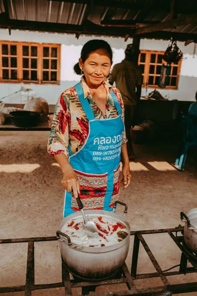 A Thai woman cooking and stirring a pot while looking and smiling at the camera.