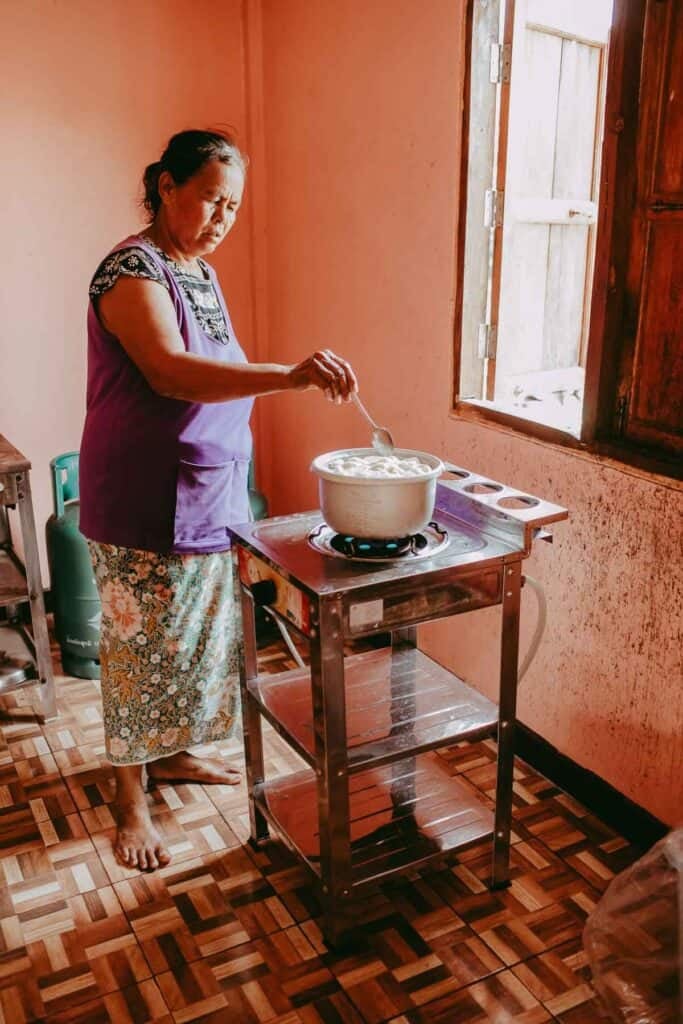 A Thai woman cooking and stirring a pot on her small stove inside a house. 