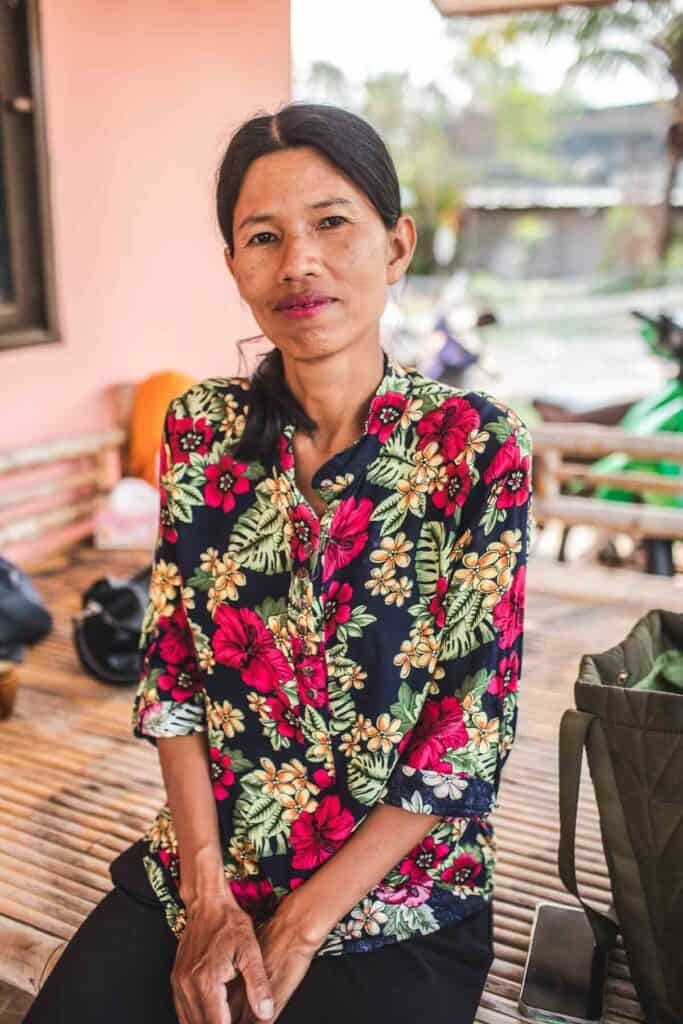 A Thai woman sitting, looking, and smiling at the camera.