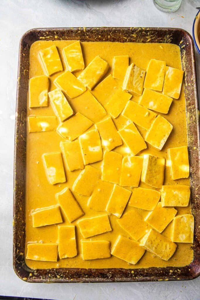 Tofu pieces on a baking tray with yellow marinating sauce. 