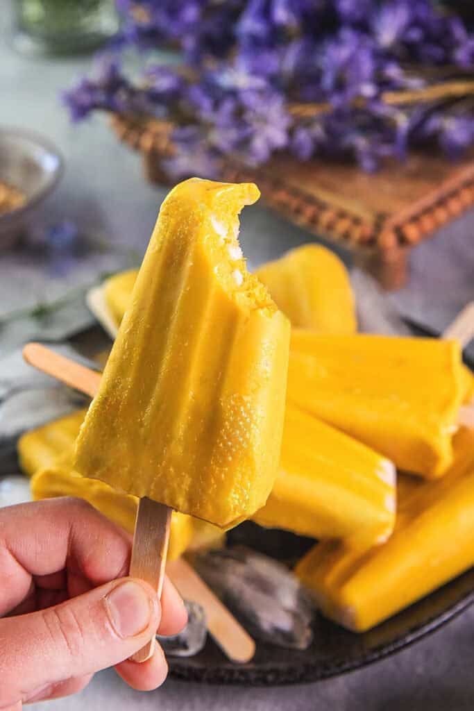 A hand holding Yellow mango Popsicles with one bite missing, with more popsicles in the background. 