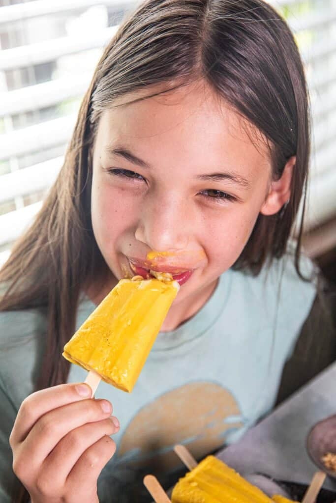 A girl eating a yellow mango popsicle. 