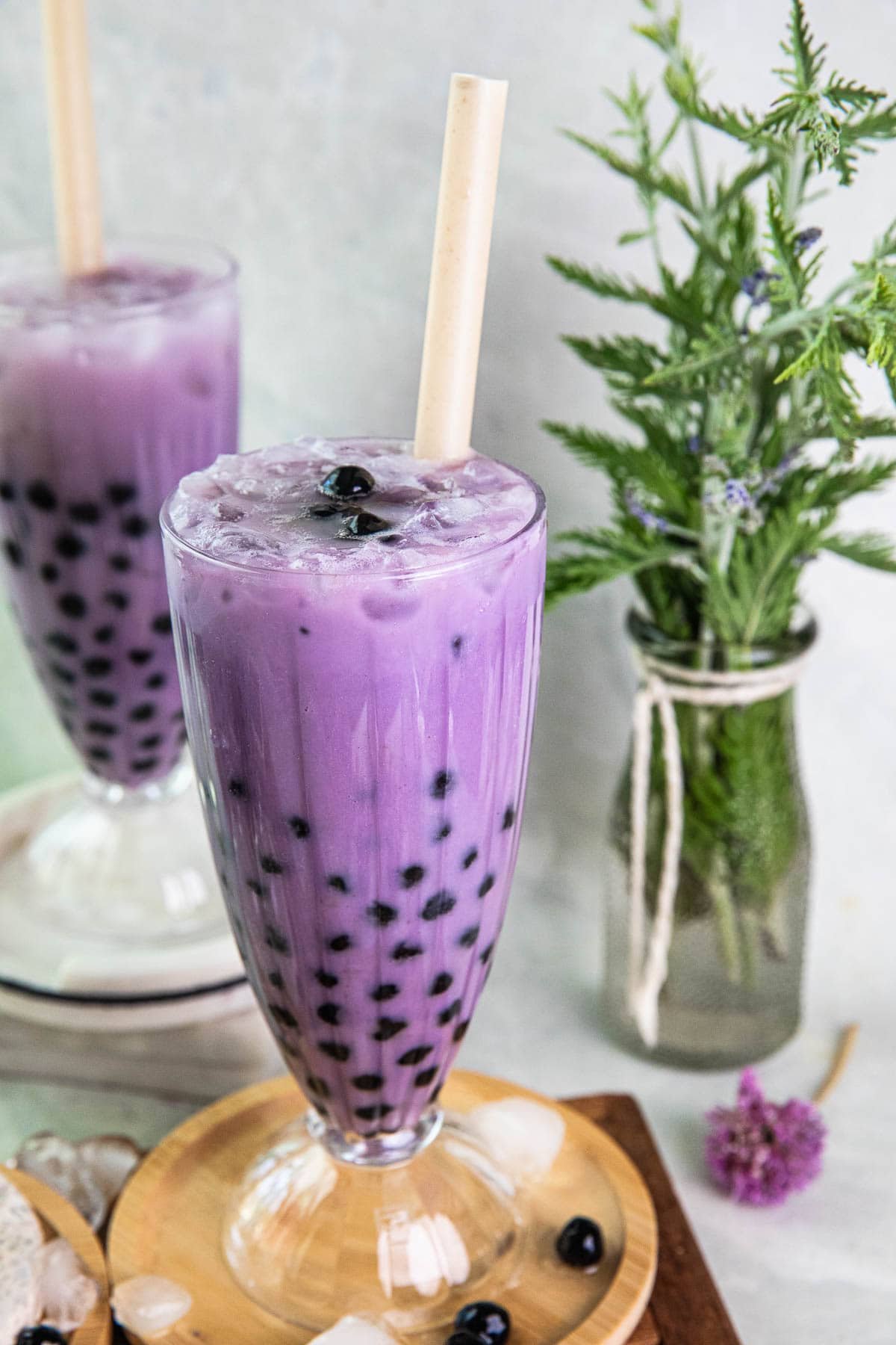 Taro boba in a tall clear glass on the table with boba and flowers in the background. 