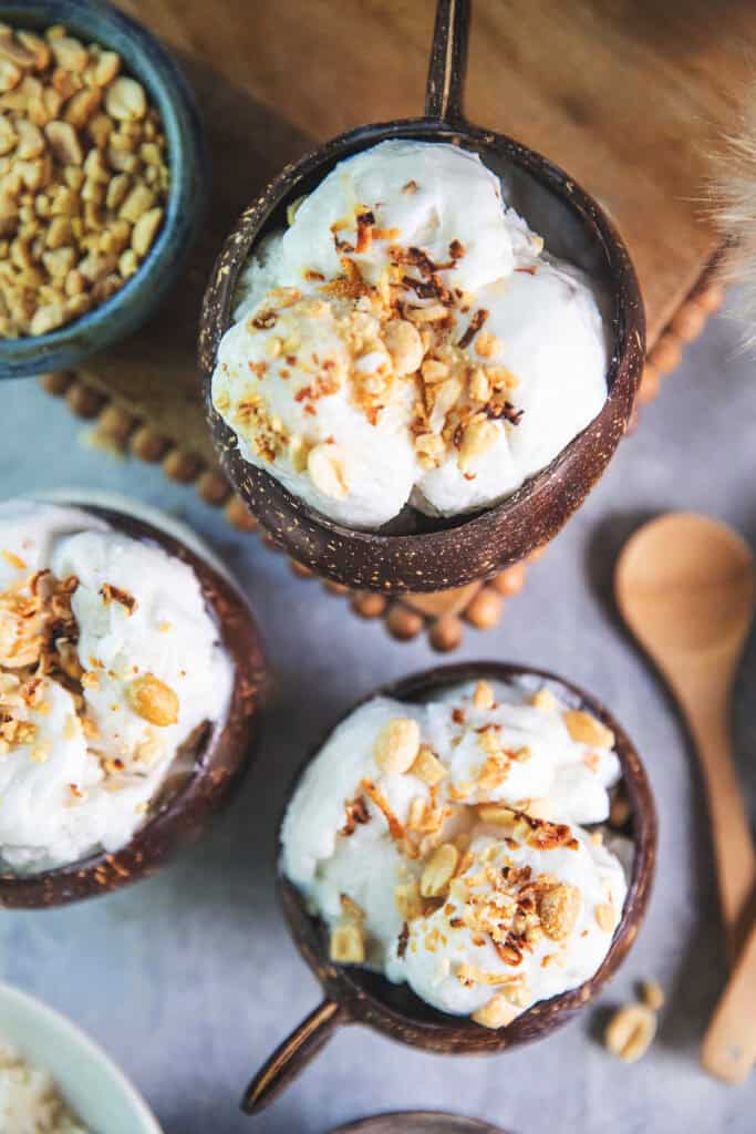 Overhead shot of Thai coconut ice cream in a coconut bowl on a wooden tray on a table with crushed peanuts on top. 
