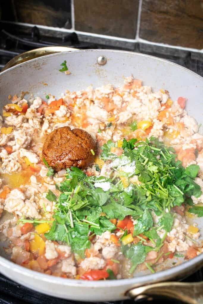 Tom Yum paste and cilantro are added on top of the meat mixture in a pan on the stove. 