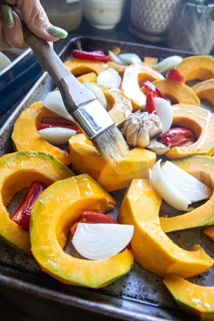 A brush with oil on Sliced pumpkin pieces, onion, peppers, and garlic on a baking sheet.