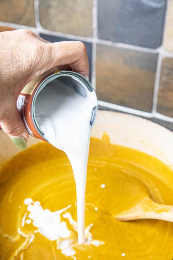A hand pouring a can of coconut milk into a pot of Thai pumpkin curry.