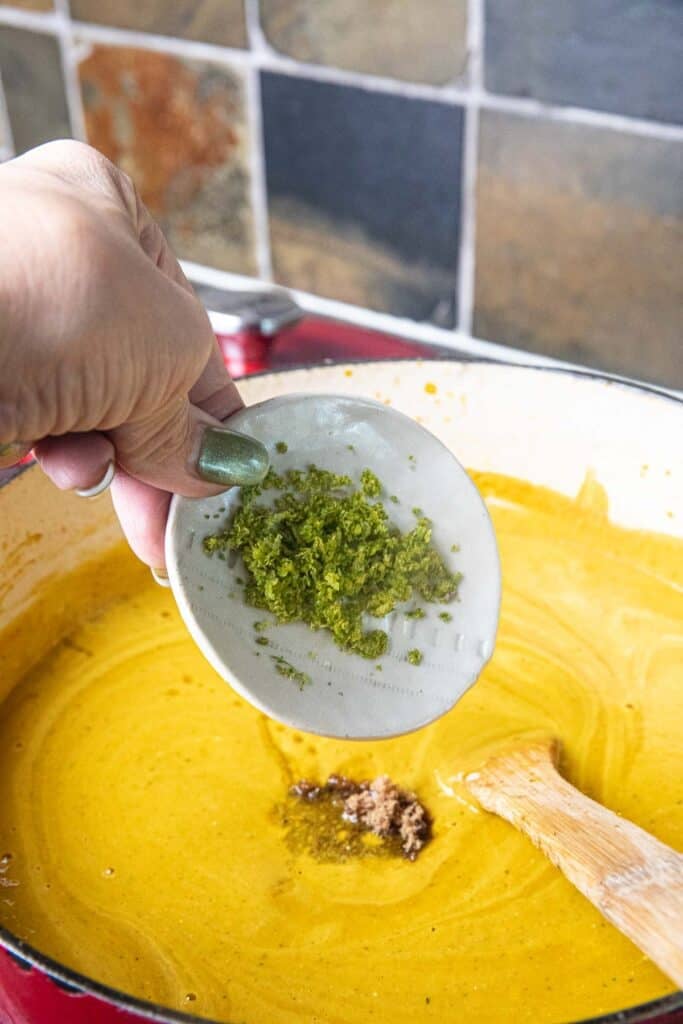 A hand pouring a small cup of lime zest into a pot of Thai curry with pumpkin.