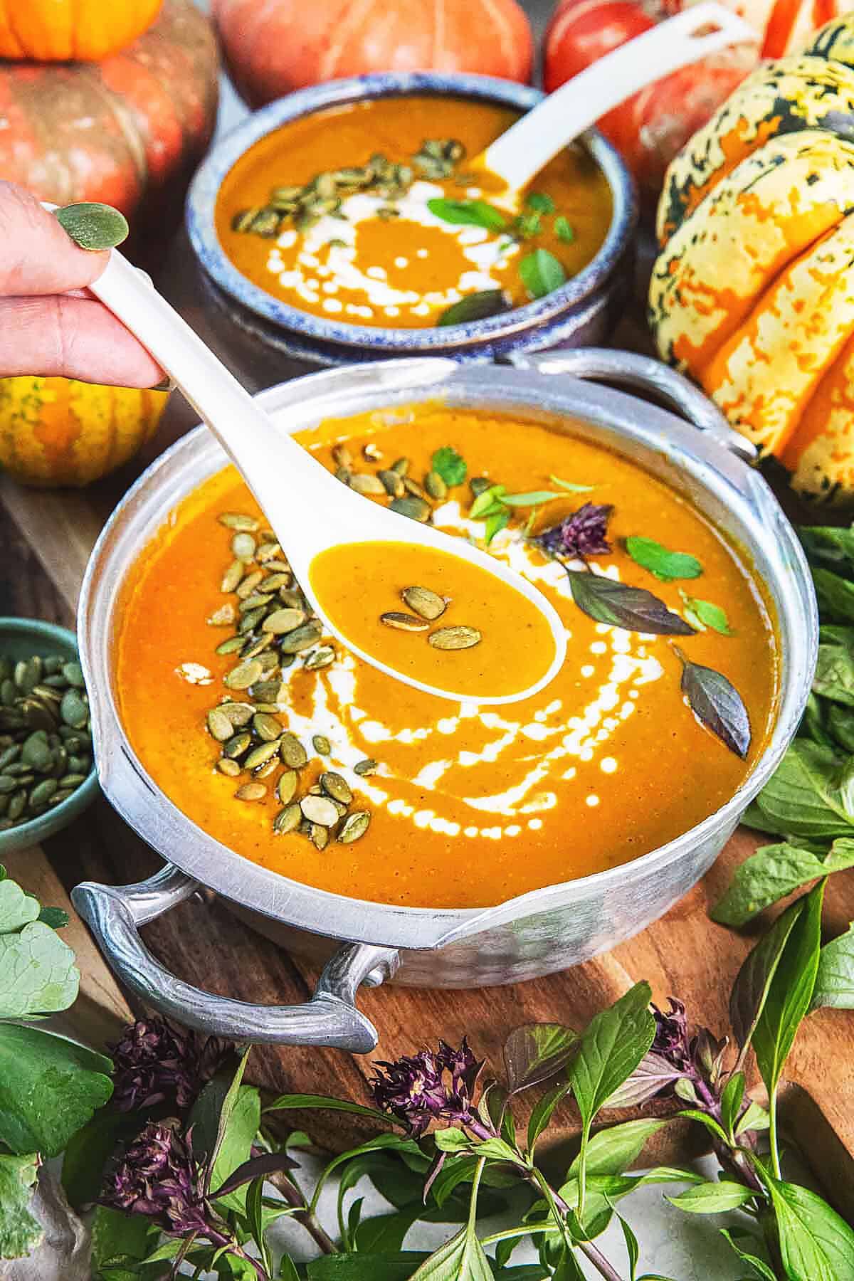 Two bowls of puree Thai pumpkin curry on a table with lots of pumpkins and basil leaves around.