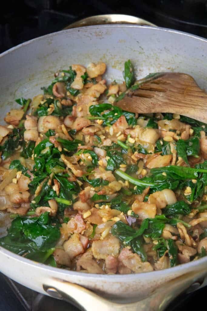 Sautéed bacon, spinach, nuts, garlic, and onion in a pan with a spatula. 
