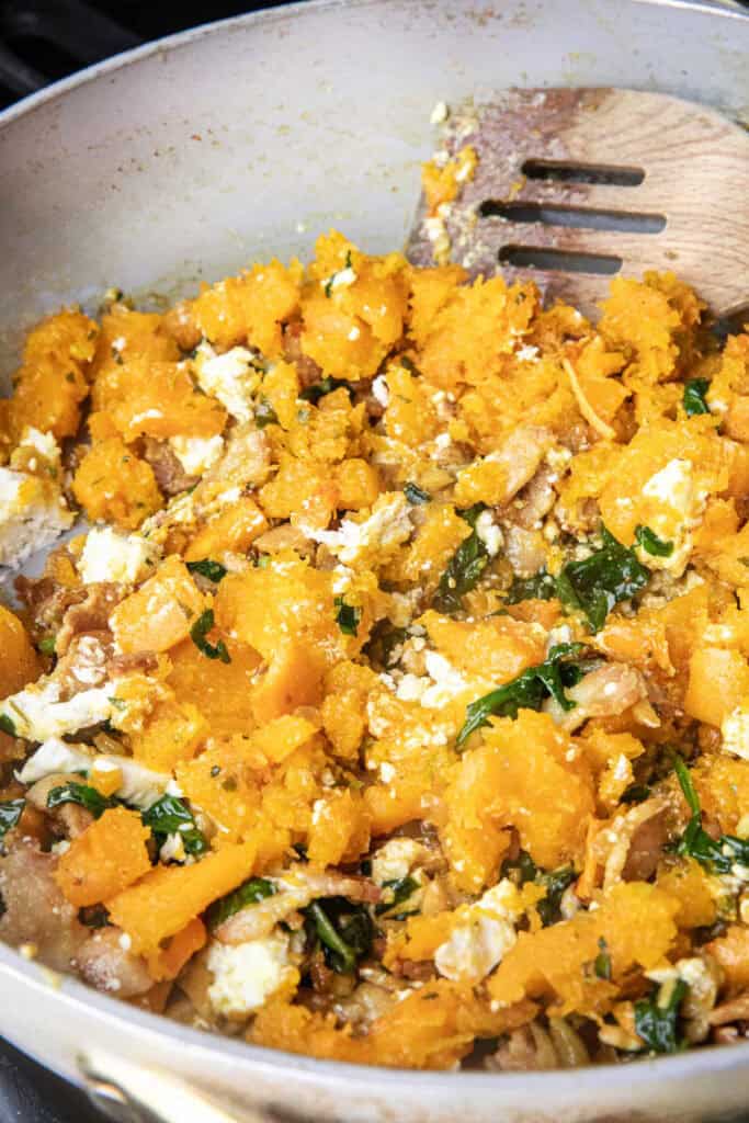 Sautéed bacon, spinach, nuts, garlic, and onion, along with creamy chunks of baked flesh, butternut squash in a pan with a spatula. 