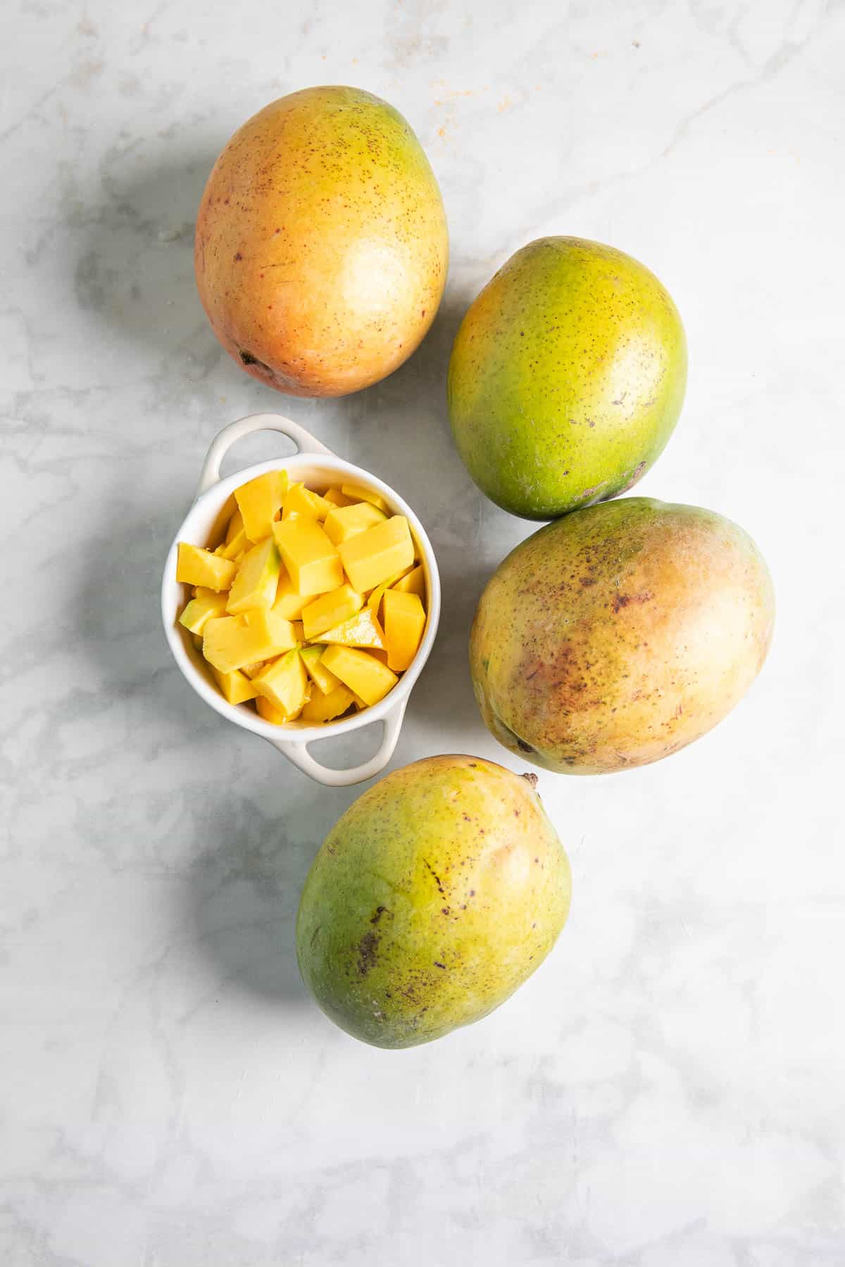 3 whole mangoes and a bowl of mango chunks on a table. 