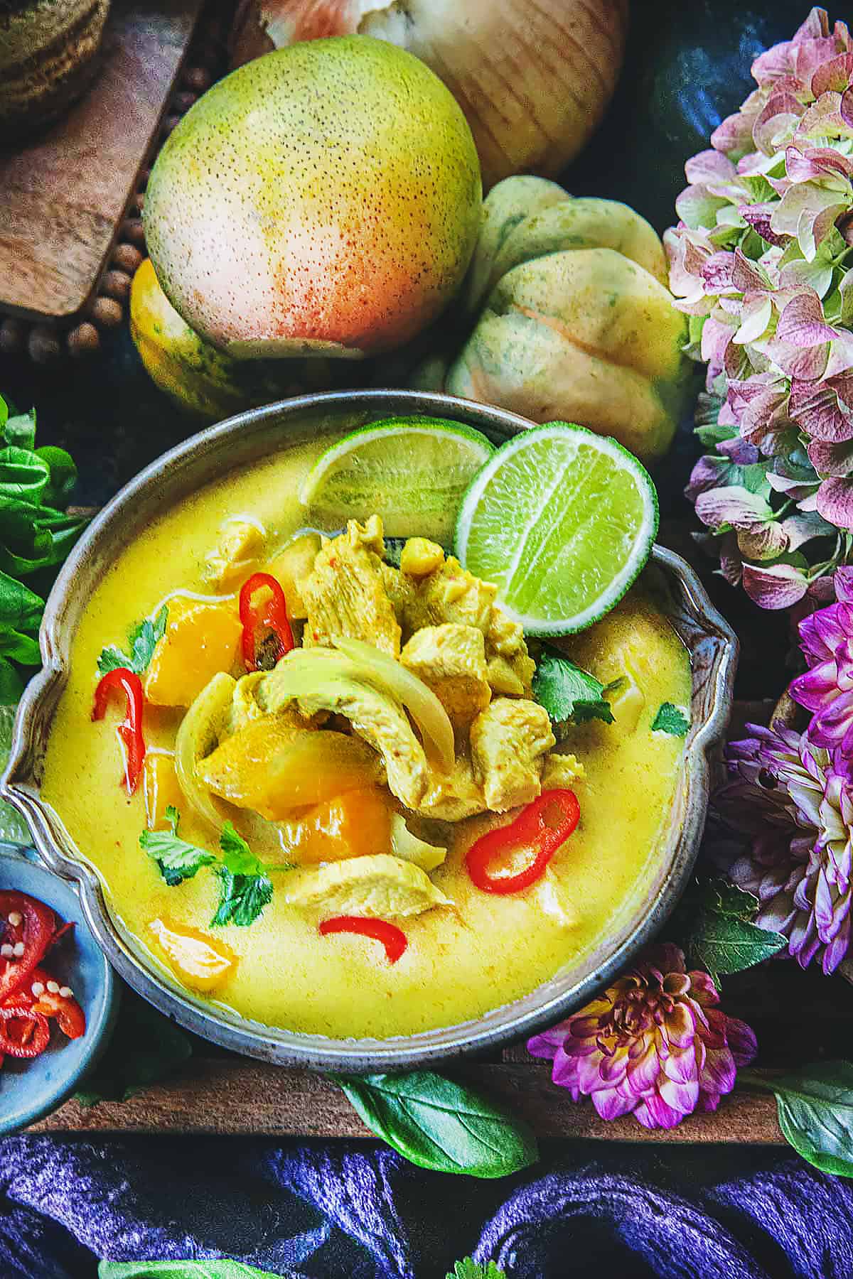 A bowl of mango curry on the table with flowers and herbs around. 
