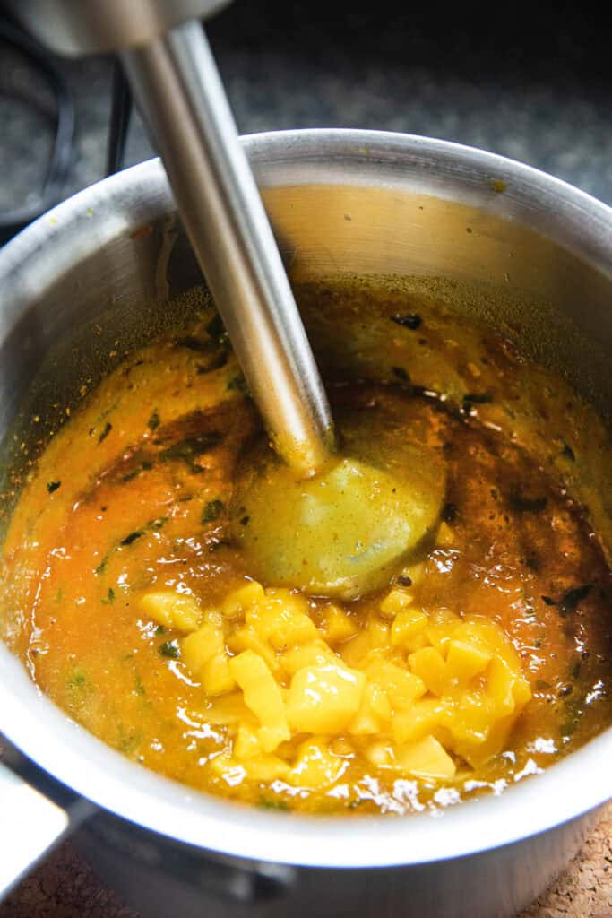 Immersion blender in a  cooked yellow mixture for mango curry sauce in a pot with mango chunks on top.