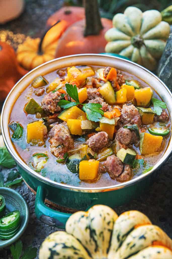 A close up of Thai pumpkin soup in a small pot and pumpkins, squashed and garnishes around it. 