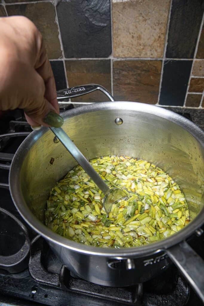 A hand stirring a pot frying fried green onions chunks in oil over a stove top.