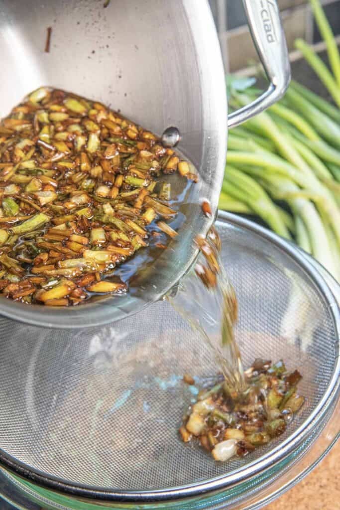 A pot pouring fried green onions and oil over a mesh strainer on top of a bowl of oil.