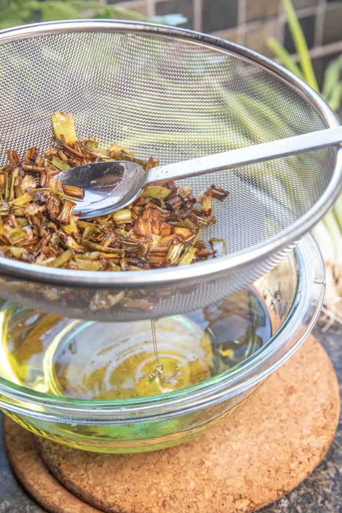 A spoon lifting fried green onion chunks over a mesh strainer on top of a bowl of oil.