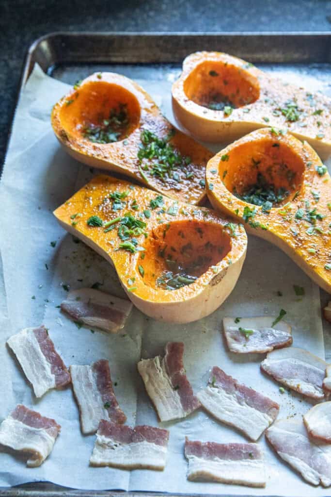 4 pieces of halved and seasoned butternut squash on a tray w bacon pieces on the side. 