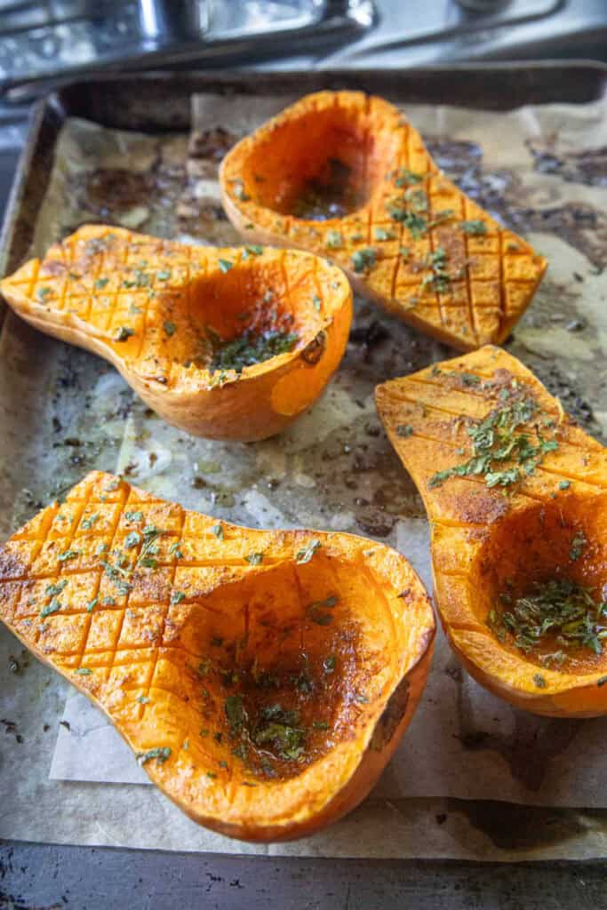 4 pieces of Baked butternut squash halves with herbs and spices on top on a baking tray. 