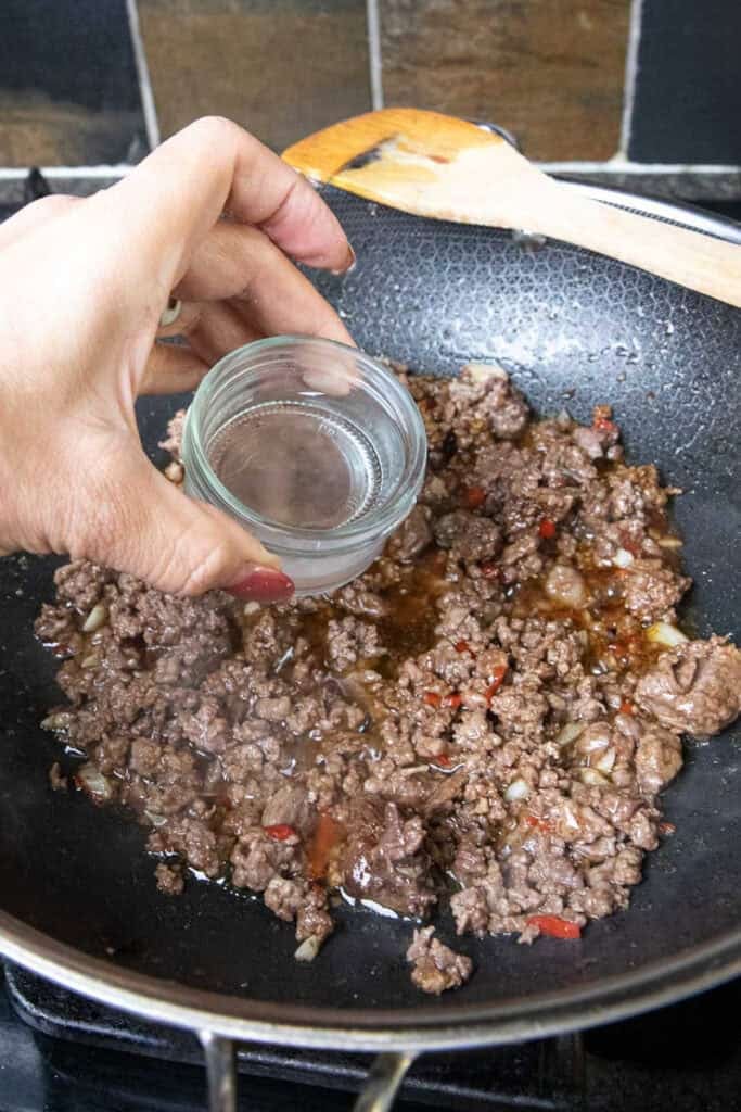 Hand pouring water into Ground beef  pad kra pao in a wok.