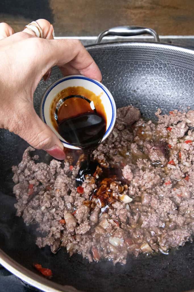 Hand pouring sauce into Ground beef  pad kra pao in a wok.