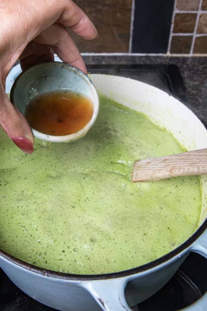 a hand pouring fish sauce into a pot of green soup on the stove. 