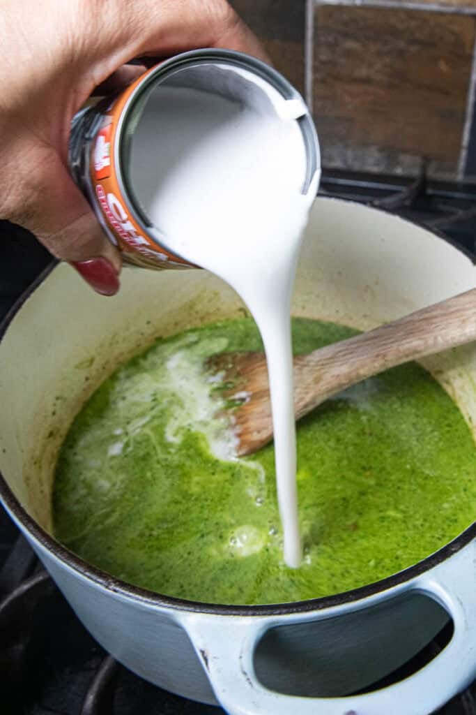 a can of coconut milk pouring into a pot of green soup on the stove. 