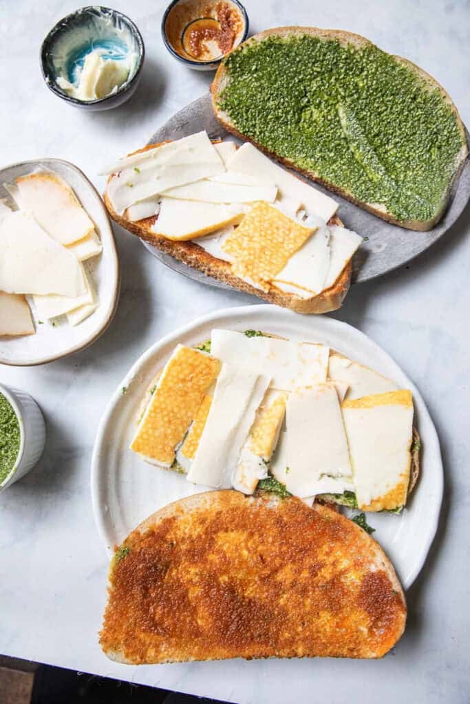 Slices of bread spread with pesto sauce, tom yum paste, and sliced cheese on top of a table.