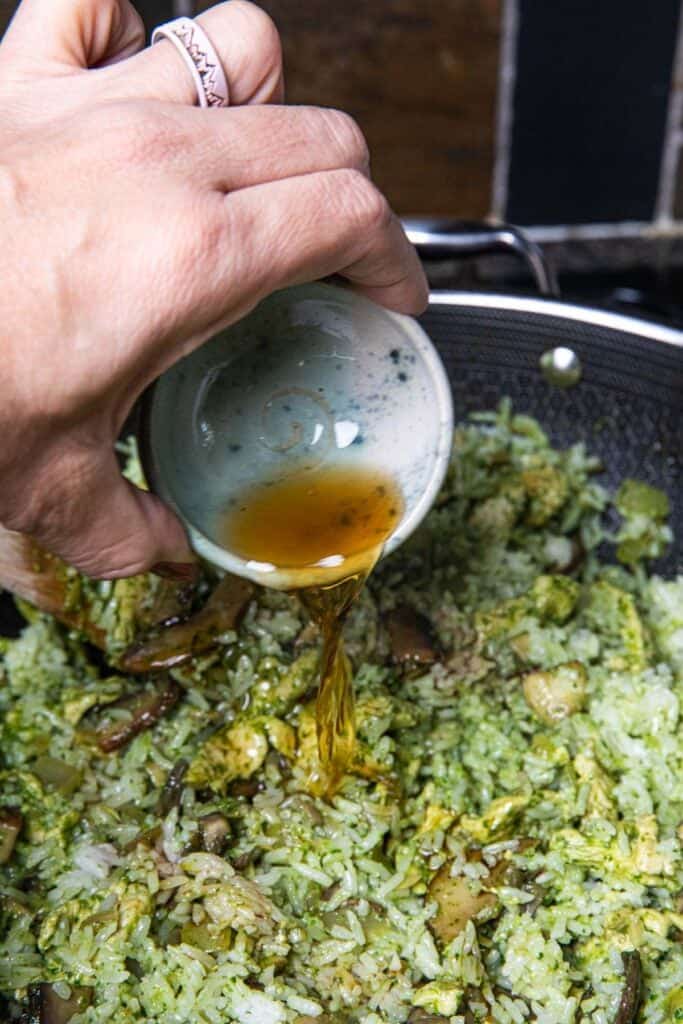 A hand pouring fish sauce into green chicken pesto fried rice in a wok.