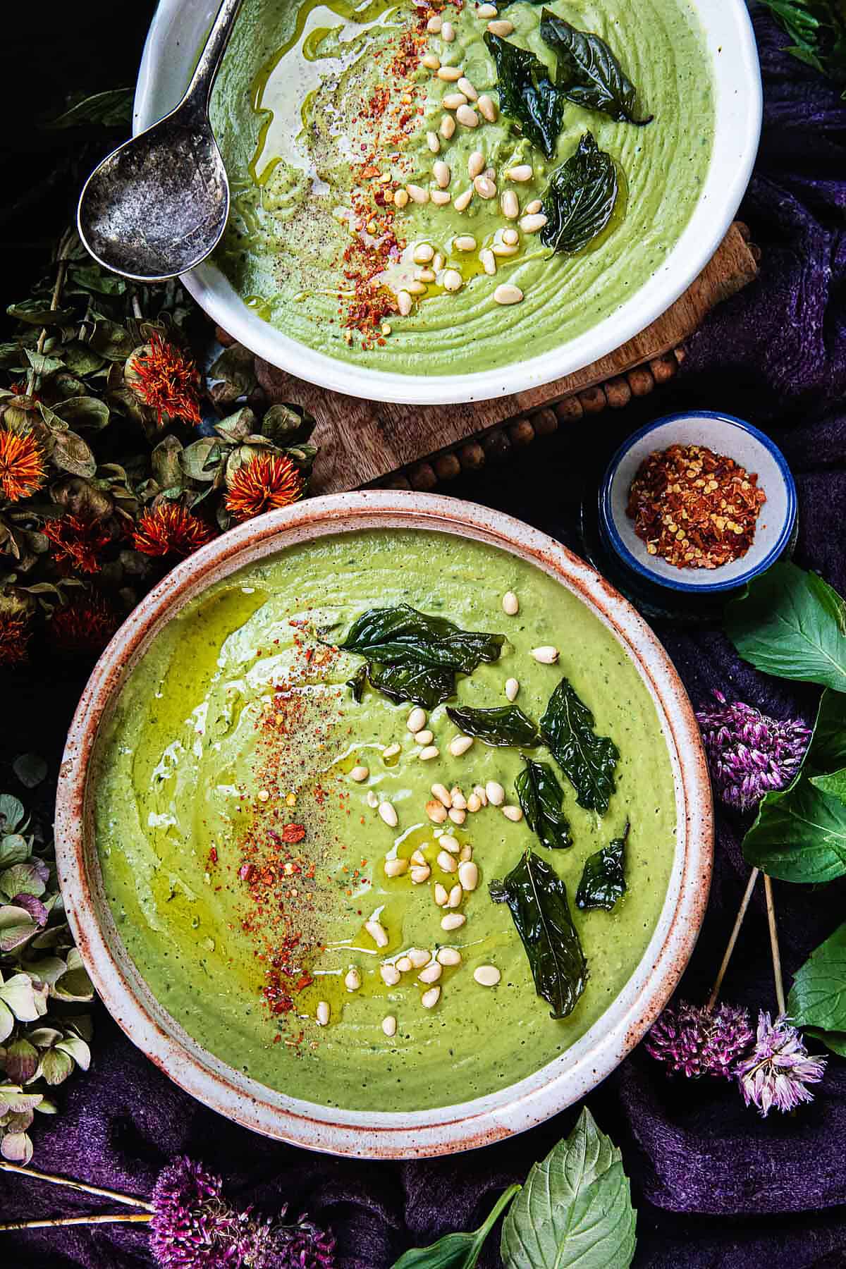 thai basil pesto soup in 2 bowls with garnishes on top surrounded by flowers and basil. 