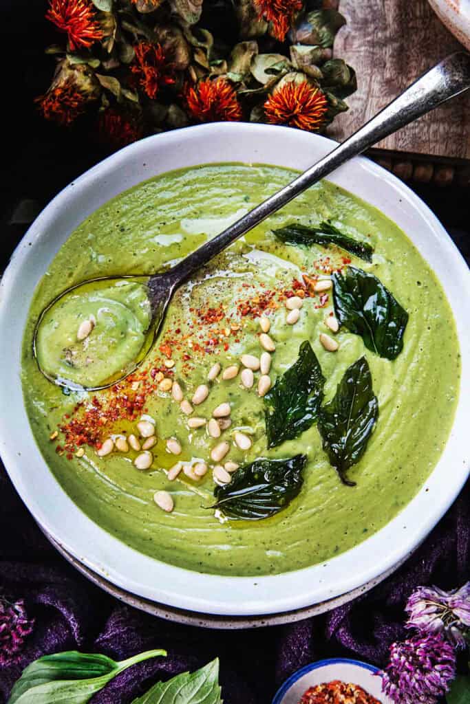Creamy Thai basil pesto soup in a bowl with a spoon, pine nuts, and fried Thai basil leaves garnished on top. 