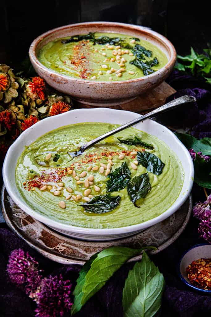 2 bowls of creamy Thai basil pesto soup in a bowl with a spoon, pine nuts, and fried Thai basil leaves garnished on top. 