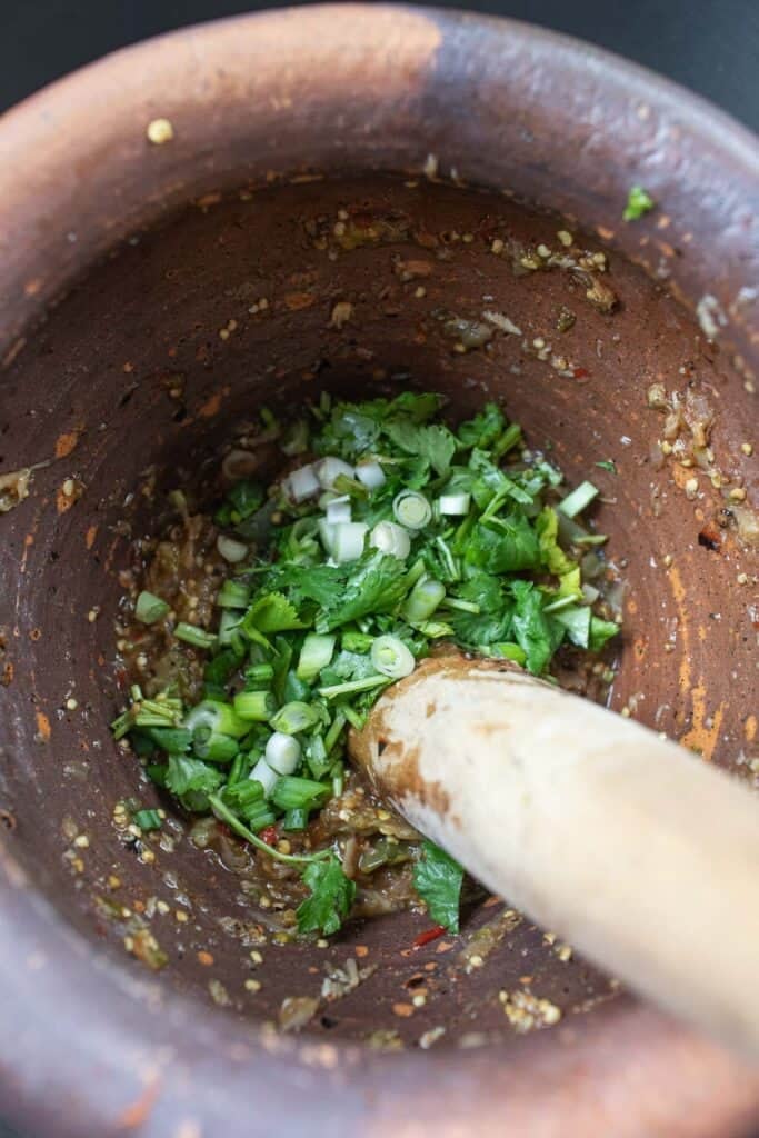 Chopped green onion and cilantro in a Thai roasted eggplant dib in a mortar with a pestle inside. 