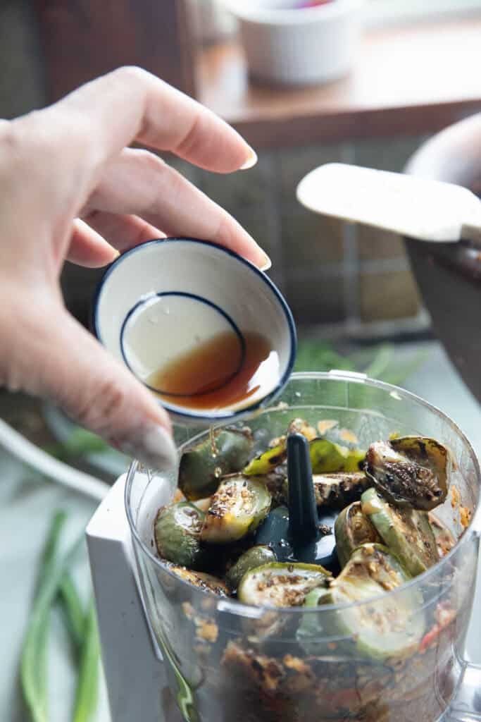 A hand pouring fish sauce into a blender with Thai eggplant dip ingredients. 