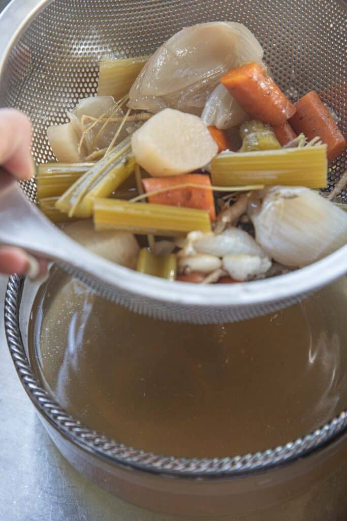 Cooked vegetables in a sieve strainer over stock in a glass bowl in the bottom.