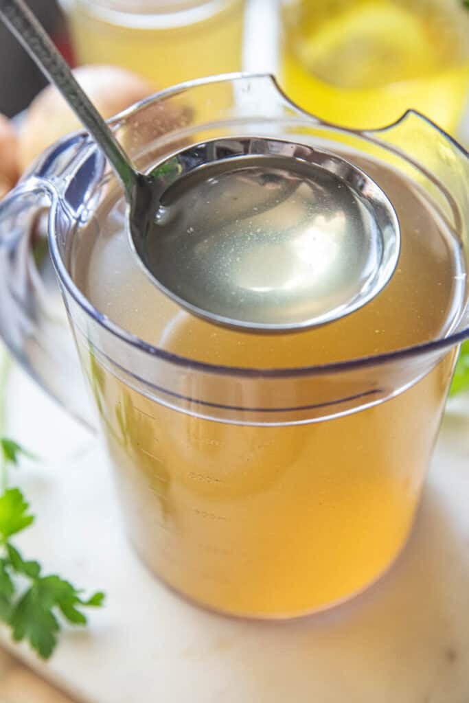 Thai Vegetable Stock in a glass jar, with a ladle in the center, on a table surrounded by herbs.
