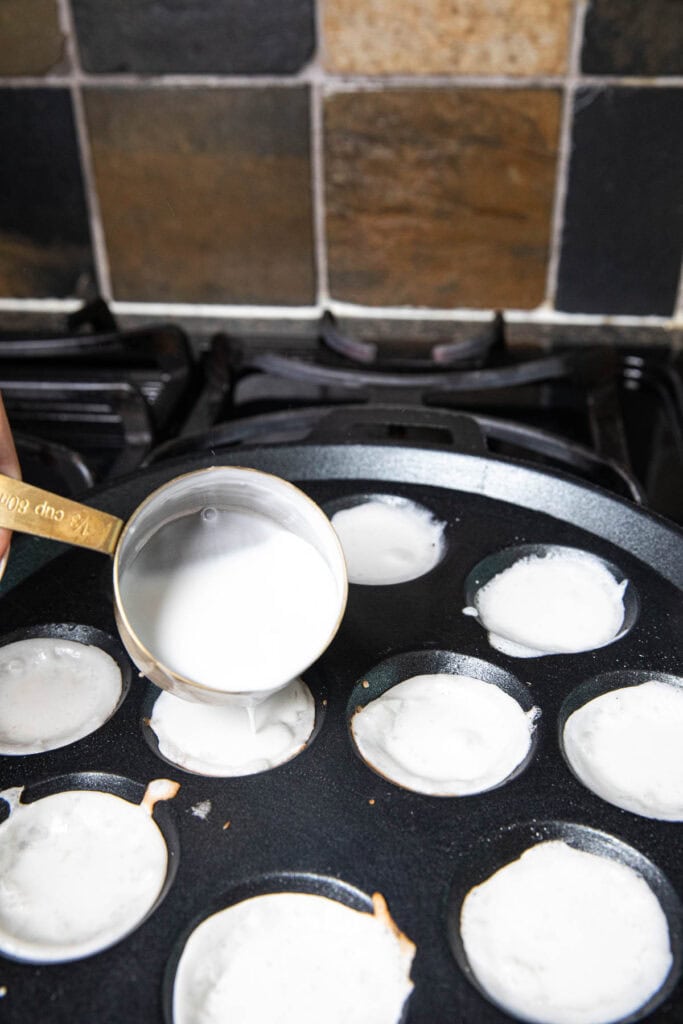Measuring cup pouring coconut mixture on a kanom krok pan on the stove.
