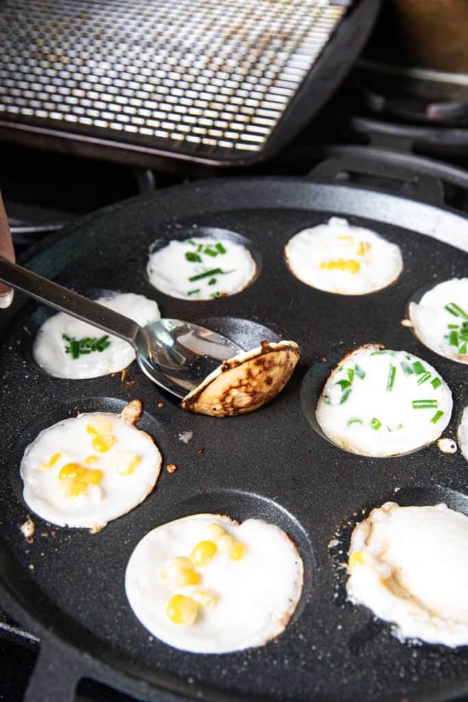 A spoon lifting thai coconut pancakes kanom krok from a pancake pans on the stove.