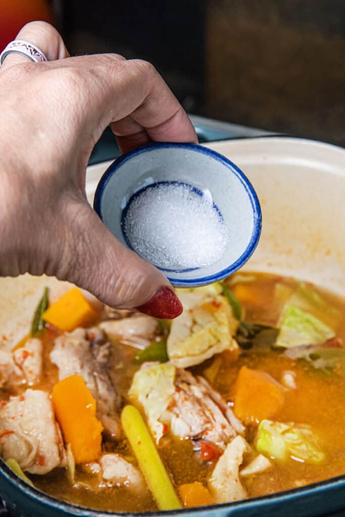 Fingers holding MSG in a small bowl pouring into a Gaeng Om in a pot.