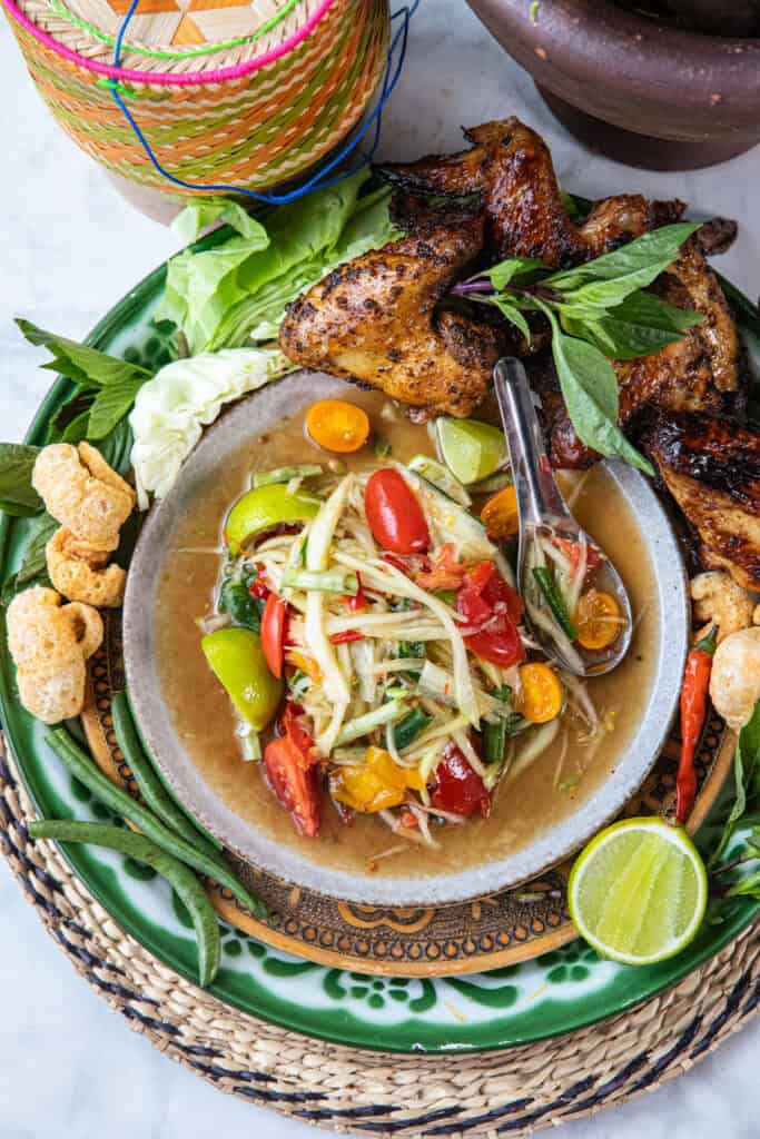A plate of Som Tum Pu Pla Ra with chicken wings, veggies, pork rind and sticky rice around it.