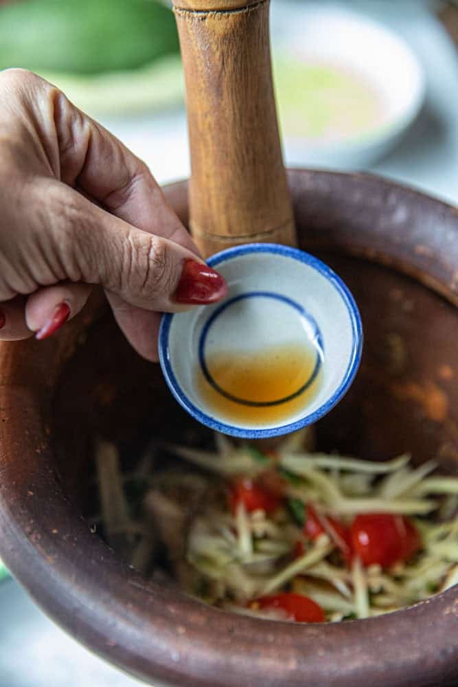 Hand holding a small bowl of fish sauce pouring into a mortar with papaya salad.