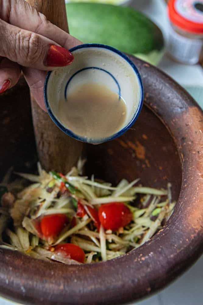 Hand holding a small bowl of pla ra sauce pouring into a mortar with papaya salad.
