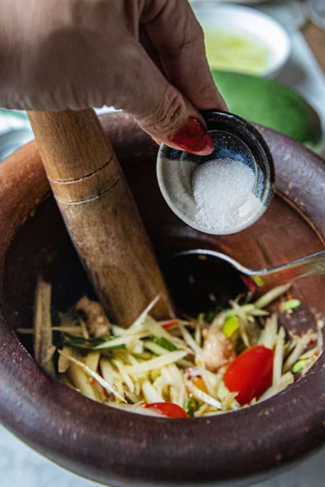 Fingers holding MSG in a small bowl pouring into a Som Tum salad in a mortar.