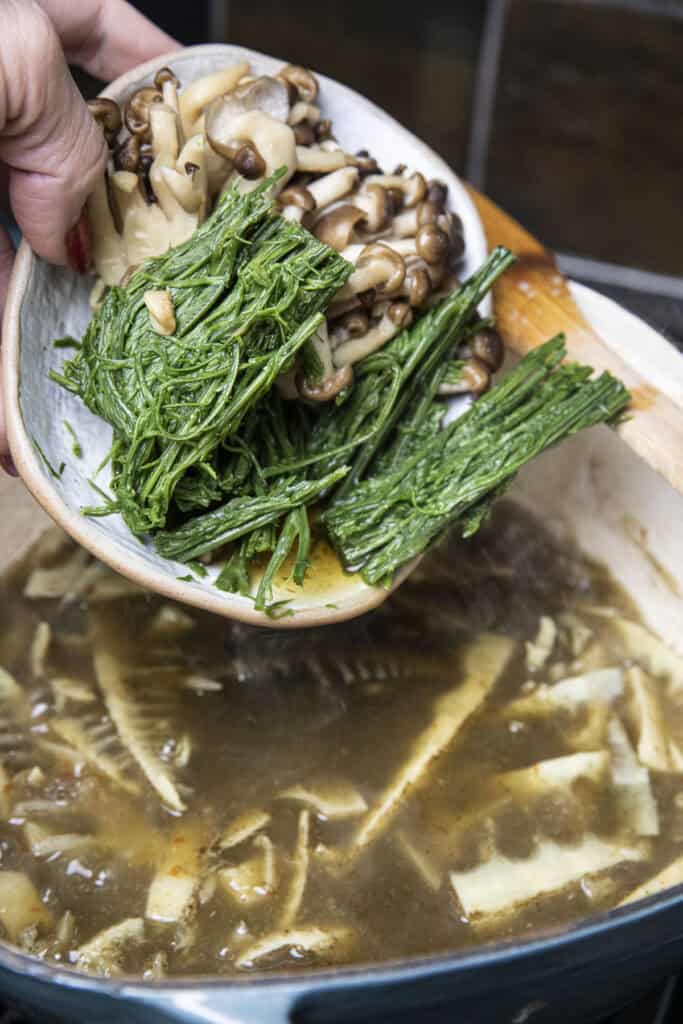 Cha Om herb and mushroom in a small plate pouring in to a pot of gaeng nor mai soup.