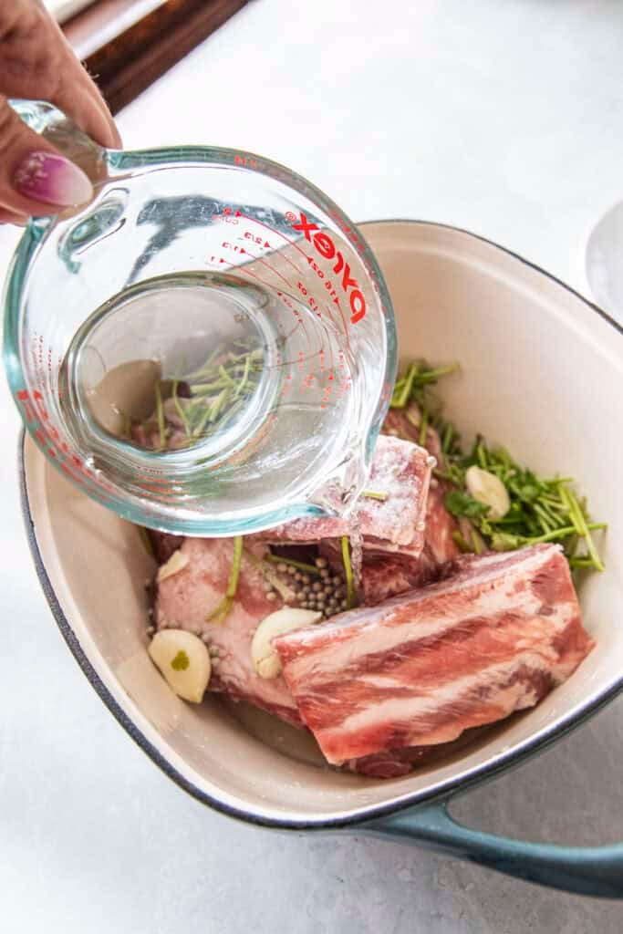 water pouring into a pot of pork ribs and ingredients for making leng Saap broth.