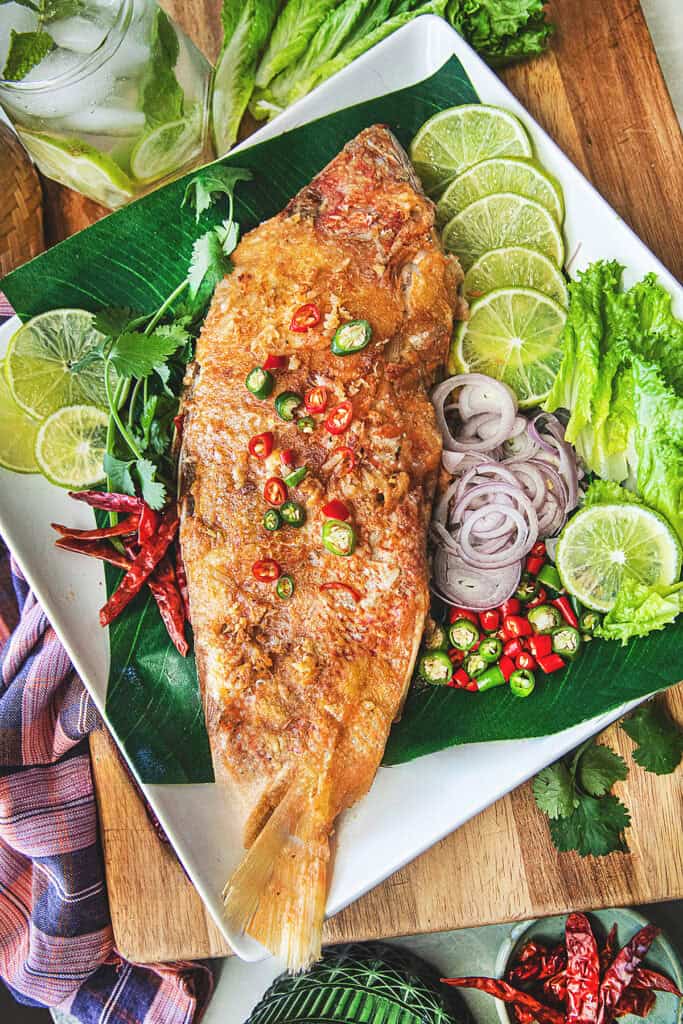 Crispy fried Pla Som topped with sliced Thai chilies, served with lime wedges and fresh herbs on a banana leaf.