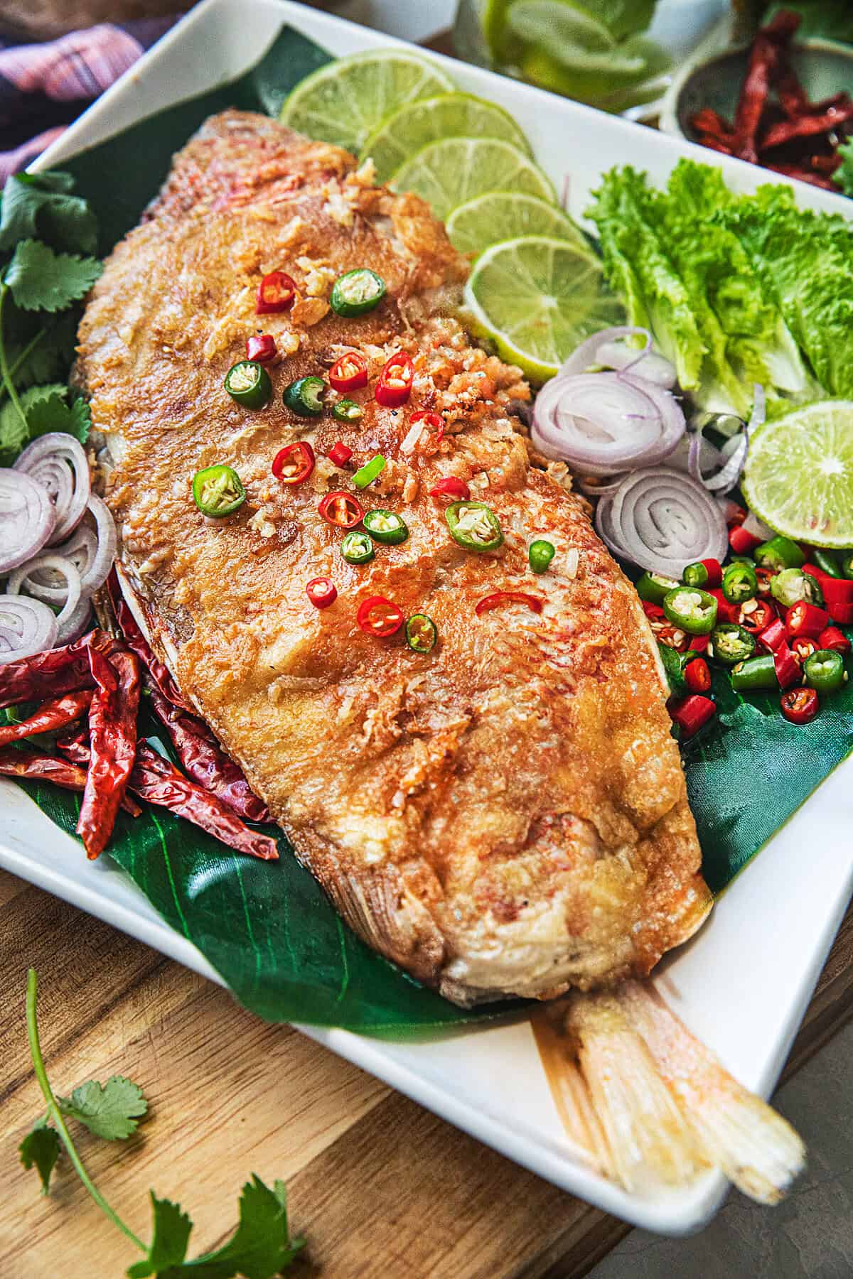 Crispy fried sour fish Pla Som topped with sliced Thai chilies, served with lime wedges and fresh herbs on a banana leaf.