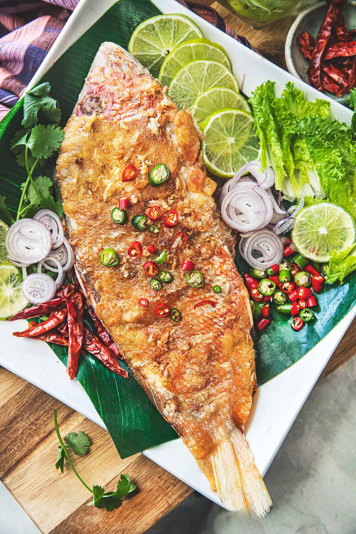 Golden pan fried Pla Som served with shallots, fresh chilies, lettuce, and lime slices on a white platter.