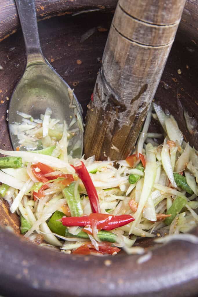 mixing thai papaya salad in clay mortar with a spoon and pestle.